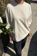 A person outdoors wearing an oversized cream-colored long-sleeve crewneck sweatshirt and dark pants, standing on a concrete sidewalk next to a planter with white flowers.