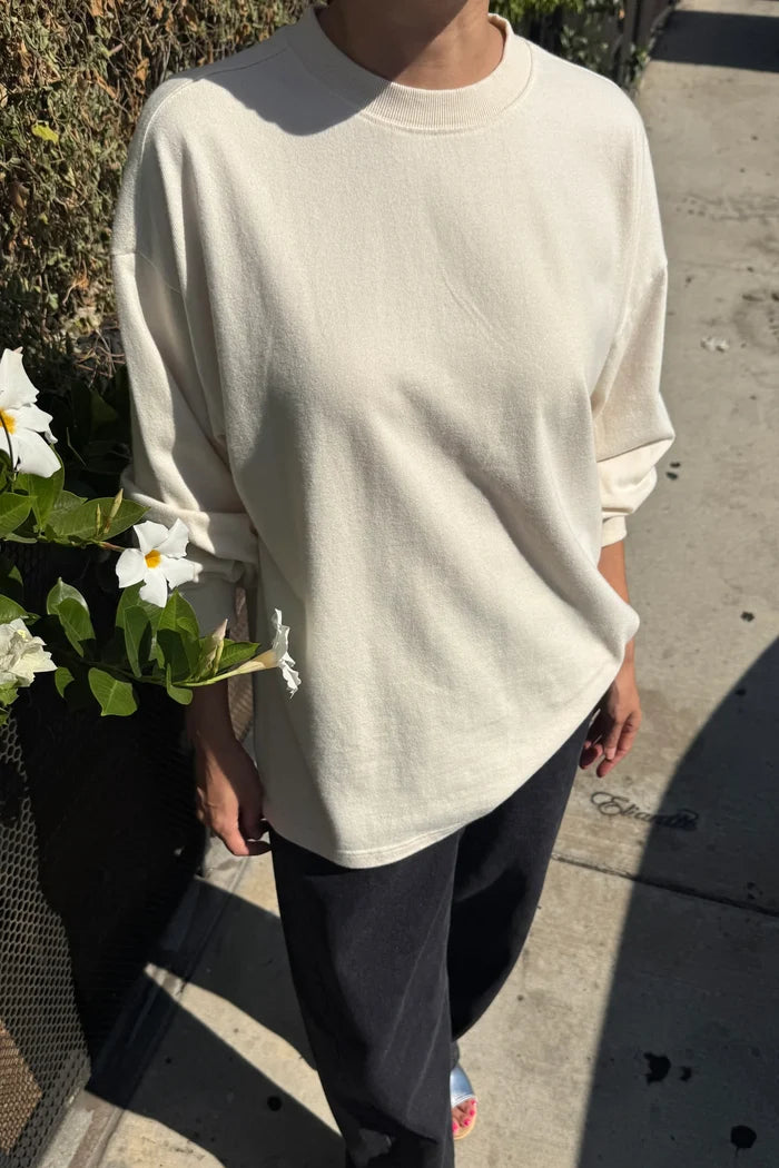 A person outdoors wearing an oversized cream-colored long-sleeve crewneck sweatshirt and dark pants, standing on a concrete sidewalk next to a planter with white flowers.