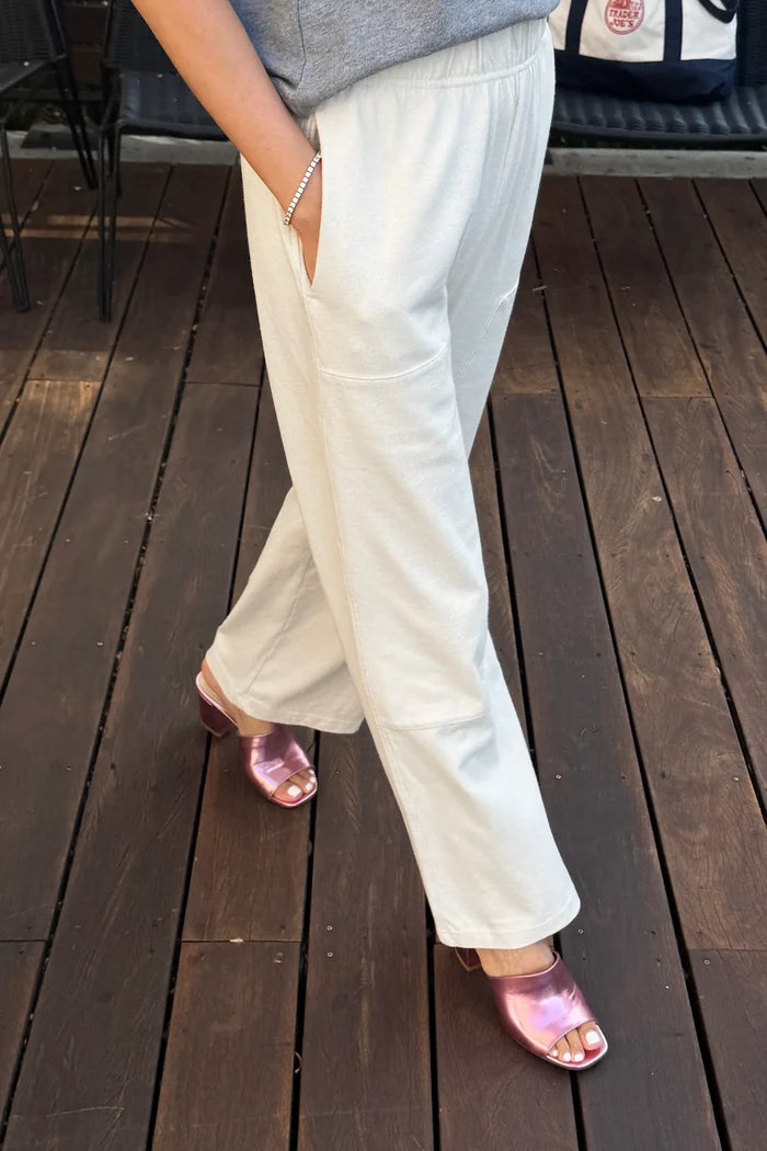 A person wearing off-white wide-leg pants with side pockets, paired with a gray top and metallic pink heeled sandals, standing on a dark wooden deck.