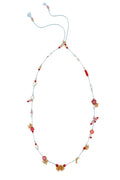 A delicate beaded necklace with a light blue cord, featuring red and white beads, small floral motifs, and gold-toned decorative accents, laid flat on a white textured background.