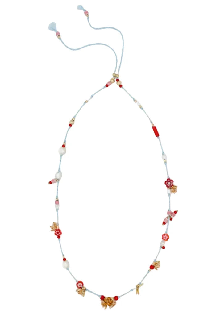 A delicate beaded necklace with a light blue cord, featuring red and white beads, small floral motifs, and gold-toned decorative accents, laid flat on a white textured background.