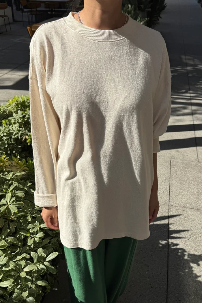 A person wearing an oversized cream-colored long-sleeve tee and green pants, standing outdoors on a paved walkway with plants and dappled sunlight.