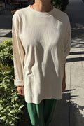 A person wearing an oversized cream-colored long-sleeve tee and green pants, standing outdoors on a paved walkway with plants and dappled sunlight.