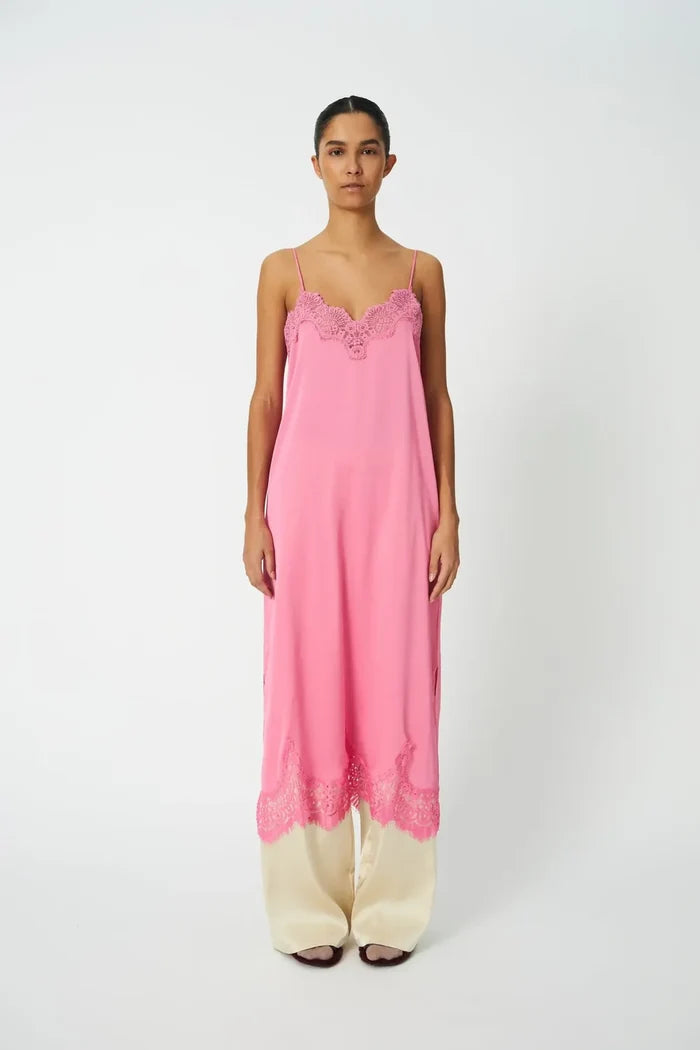 A woman stands against a plain light gray studio background wearing a long pink slip-style nightgown with thin straps and lace trim at the neckline and hem, paired with wide-leg cream pants.