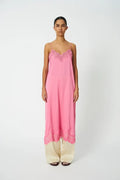 A woman stands against a plain light gray studio background wearing a long pink slip-style nightgown with thin straps and lace trim at the neckline and hem, paired with wide-leg cream pants.