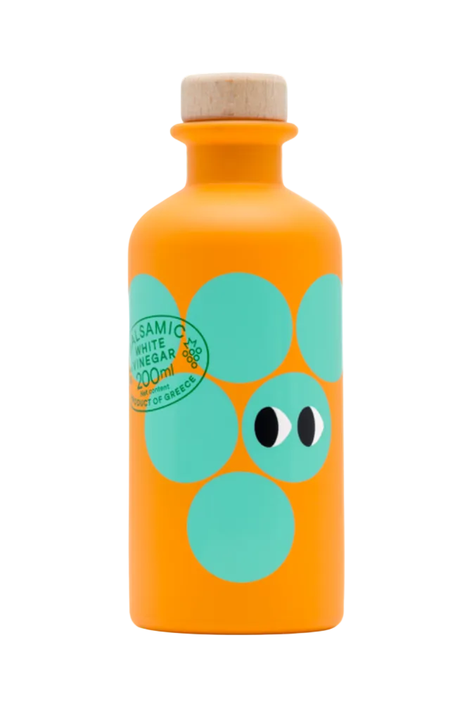 An orange bottle of Greek balsamic vinegar with a cork stopper, decorated with large teal circles and a cartoon face, on a white background.