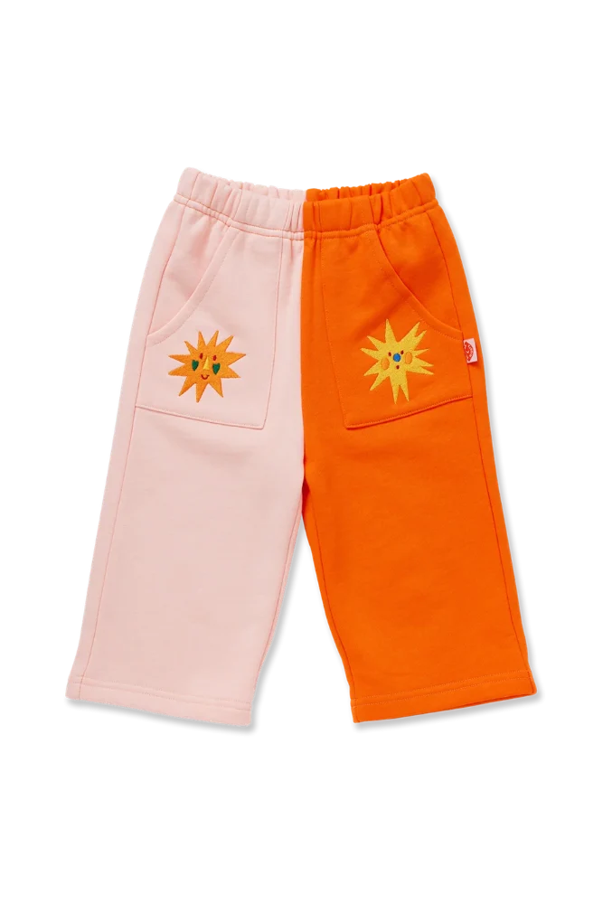 A pair of children's sweatpants laid flat on a white background, featuring a split design with one leg in light pink and the other in bright orange. Each leg has a front patch pocket with a yellow sun patch embroidered on it.