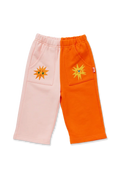 A pair of children's sweatpants laid flat on a white background, featuring a split design with one leg in light pink and the other in bright orange. Each leg has a front patch pocket with a yellow sun patch embroidered on it.