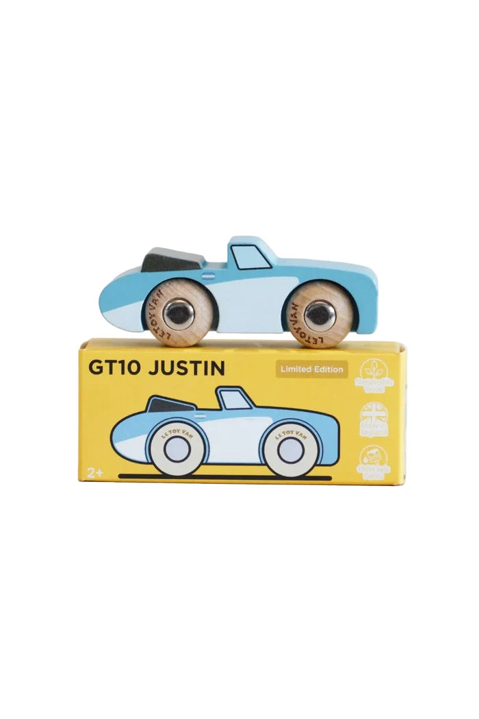 A light blue and white wooden toy race car with natural wood wheels sits on top of its yellow 'GT10 Justin Limited Edition' storage box. The box features a graphic of the toy car and age suitability markings of 2+.